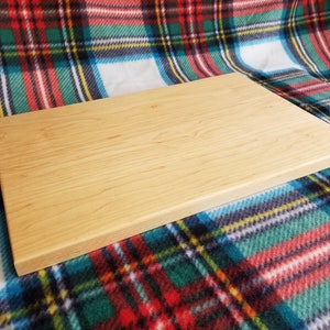 May include: A rectangular wooden cutting board made of light wood with a smooth surface. The board is lying on a red, green, and blue plaid fabric.
