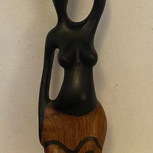 Pode incluir: Escultura de madeira de uma mulher a carregar um pote na cabe&ccedil;a. A escultura &eacute; pintada de preto e castanho com um padr&atilde;o de gr&atilde;o de madeira.