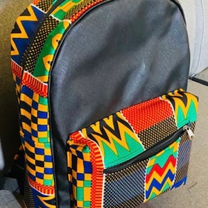 May include: A black leather backpack with a colorful African print design. The backpack has a large main compartment and a smaller front pocket. The print features geometric shapes in yellow, red, green, blue, and black.