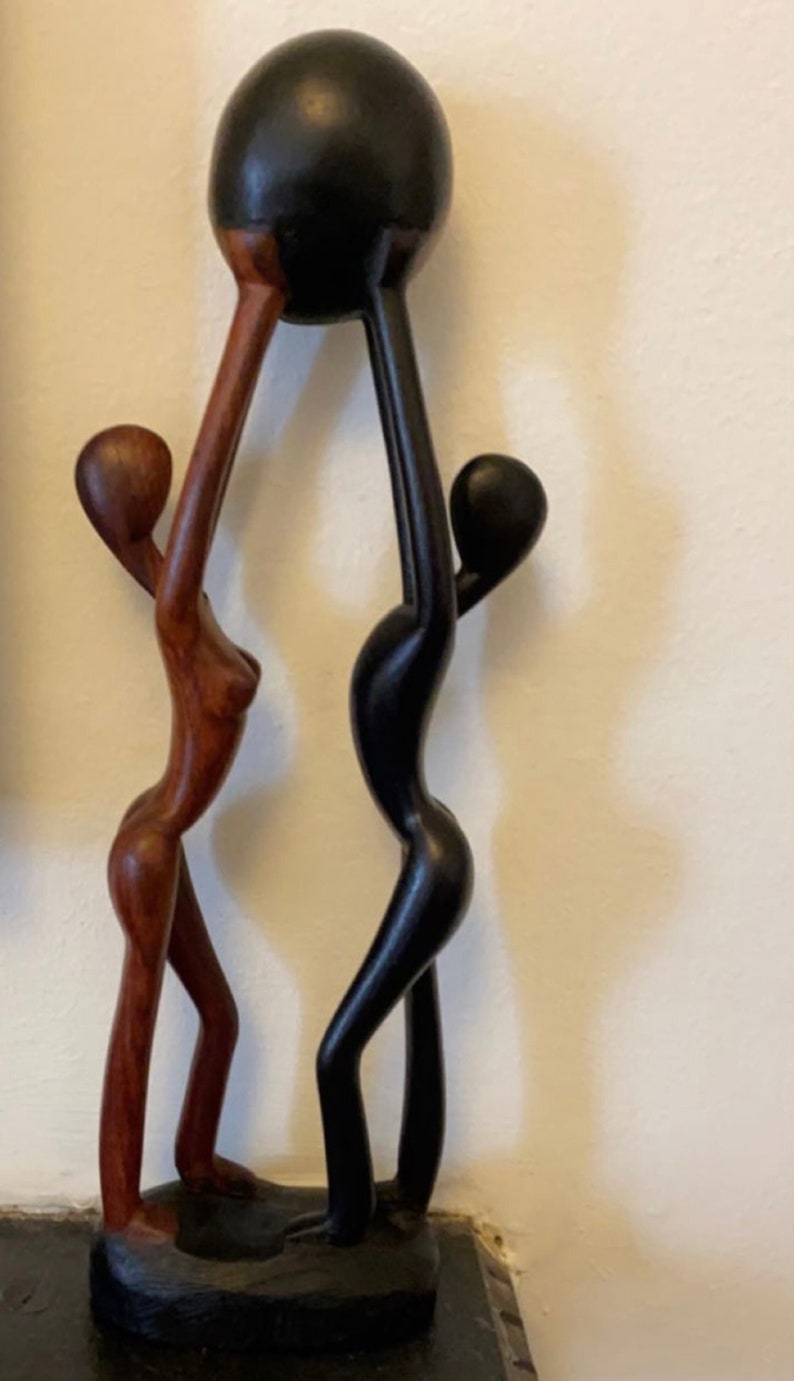 Pode incluir: Uma escultura de madeira de duas figuras segurando uma esfera preta acima de suas cabe&ccedil;as. A figura &agrave; esquerda &eacute; marrom e a figura &agrave; direita &eacute; preta.