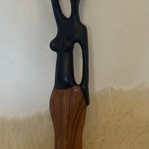 Pode incluir: Uma escultura de madeira de uma mulher carregando um pote na cabe&ccedil;a. A escultura &eacute; pintada de preto e marrom, e a mulher est&aacute; usando uma saia. A escultura est&aacute; em p&eacute; sobre uma base de madeira.