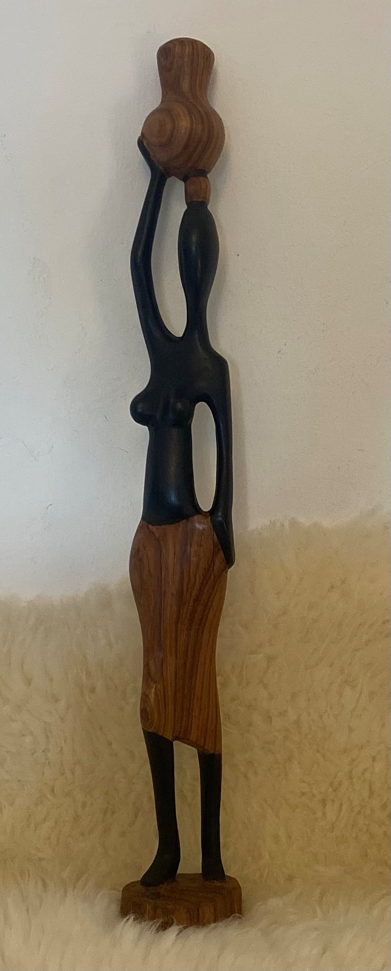 Pode incluir: Uma escultura de madeira de uma mulher carregando um pote na cabe&ccedil;a. A escultura &eacute; pintada de preto e marrom, e a mulher est&aacute; usando uma saia. A escultura est&aacute; em p&eacute; sobre uma base de madeira.
