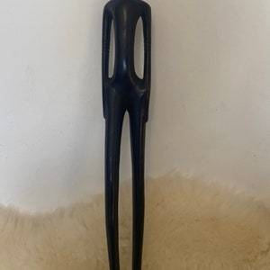 Pode incluir: Uma escultura de madeira preta e alta que representa uma pessoa com um pesco&ccedil;o longo e fino e bra&ccedil;os. A escultura est&aacute; em p&eacute; sobre uma pequena base de madeira.