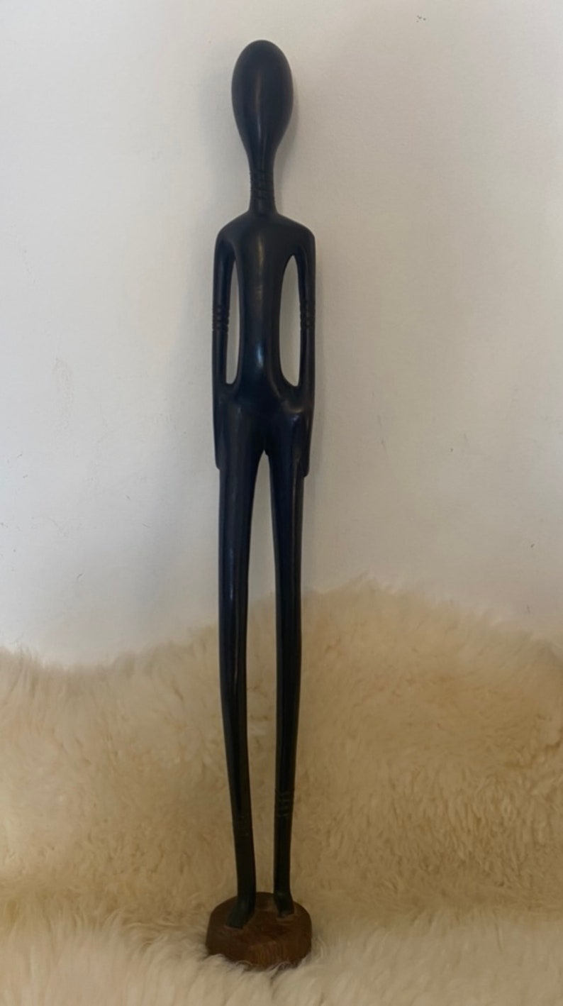 Pode incluir: Uma escultura de madeira preta e alta que representa uma pessoa com um pesco&ccedil;o longo e fino e bra&ccedil;os. A escultura est&aacute; em p&eacute; sobre uma pequena base de madeira.