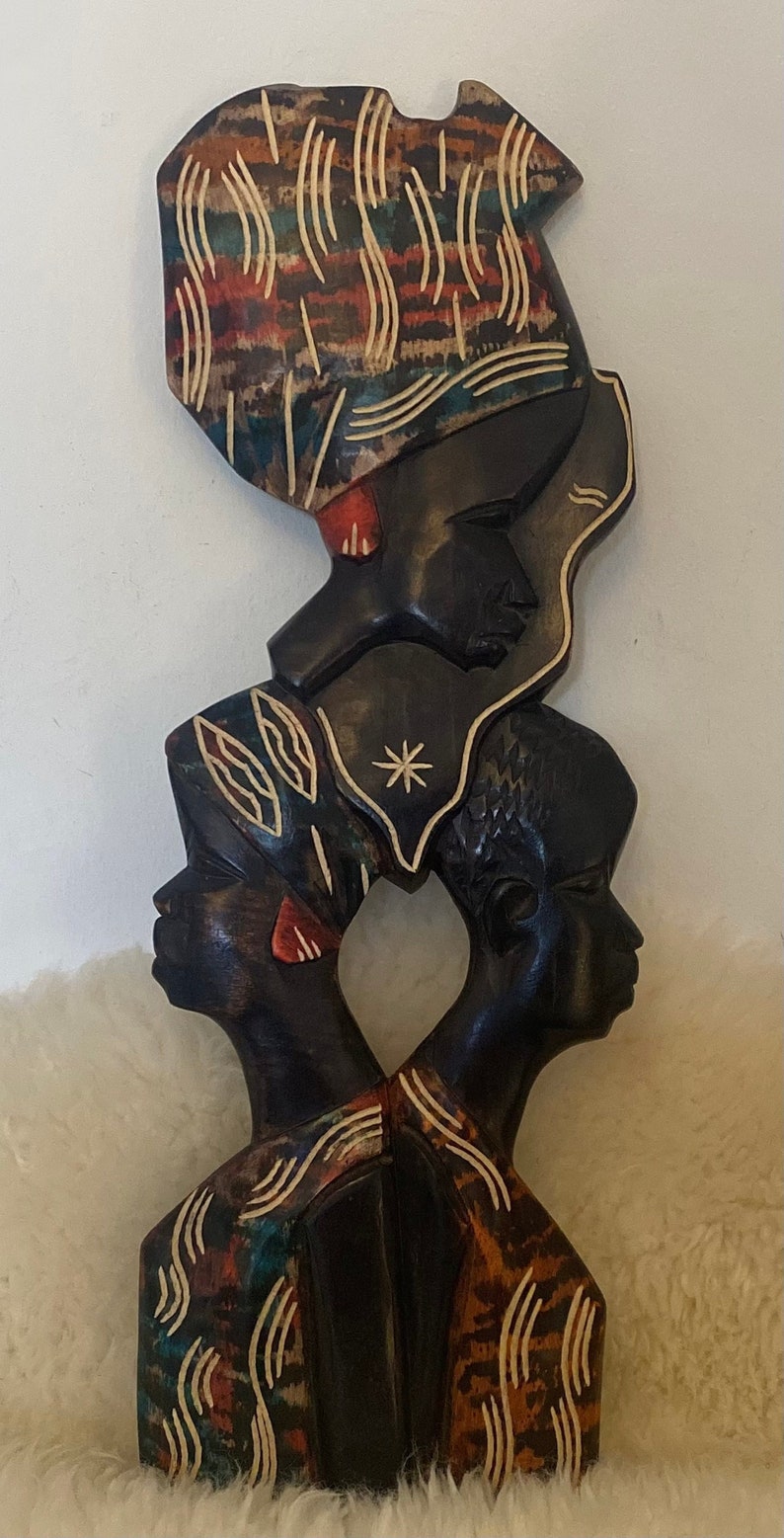 Pode incluir: Uma escultura de madeira de tr&ecirc;s figuras africanas com detalhes intrincados esculpidos. As figuras est&atilde;o empilhadas uma em cima da outra, com a figura superior usando um turbante estampado. A escultura &eacute; pintada em tons de marrom, preto, vermelho e branco.