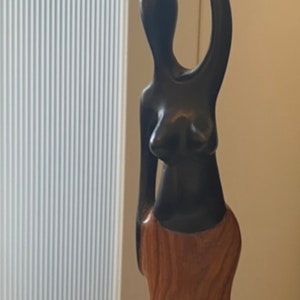 Pode incluir: Uma escultura de madeira de uma pessoa carregando um pote na cabe&ccedil;a. A escultura &eacute; feita de madeira marrom escura e marrom clara. A pessoa est&aacute; usando uma saia e tem um design simples e estilizado.