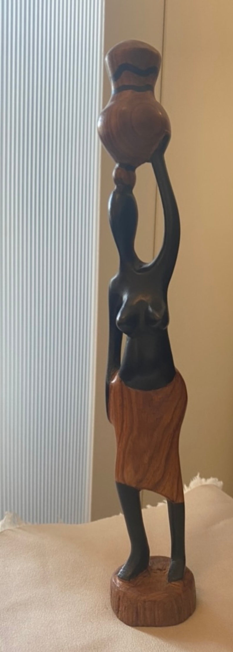 Pode incluir: Uma escultura de madeira de uma pessoa carregando um pote na cabe&ccedil;a. A escultura &eacute; feita de madeira marrom escura e marrom clara. A pessoa est&aacute; usando uma saia e tem um design simples e estilizado.