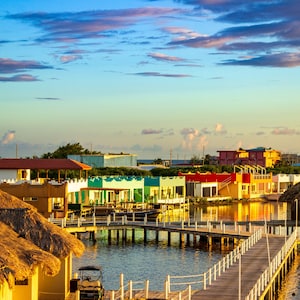 May include: A colorful row of houses on stilts over a calm body of water. The houses have thatched roofs and are connected by wooden walkways. The sky is a vibrant blue with fluffy white clouds.