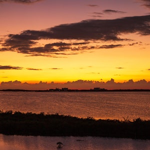 May include: A scenic sunset over a body of water with a silhouette of a distant city skyline. The sky is a vibrant orange and yellow with streaks of pink and purple clouds.