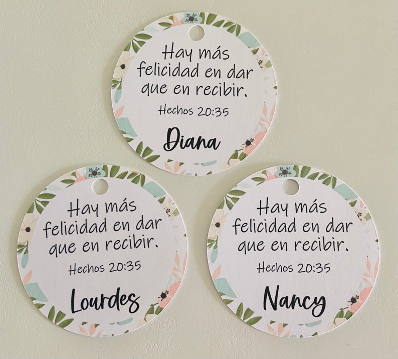 K&ouml;nnte beinhalten: Drei runde wei&szlig;e Geschenkanh&auml;nger mit Blumenmustern und gr&uuml;nen Bl&auml;ttern. Jeder Anh&auml;nger hat ein Zitat auf Spanisch, das lautet "Hay m&aacute;s felicidad en dar que en recibir." Das Zitat wird von "Hechos 20:35" gefolgt. Jeder Anh&auml;nger hat auch einen Namen: Diana, Lourdes und Nancy.