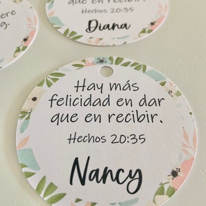 K&ouml;nnte beinhalten: Zwei runde wei&szlig;e Geschenkanh&auml;nger mit Blumenmuster. Die Anh&auml;nger haben oben ein Loch f&uuml;r eine Schnur. Der Text auf den Anh&auml;ngern lautet "Hay m&aacute;s felicidad en dar que en recibir. Hechos 20:35" und die Namen "Diana" und "Nancy".
