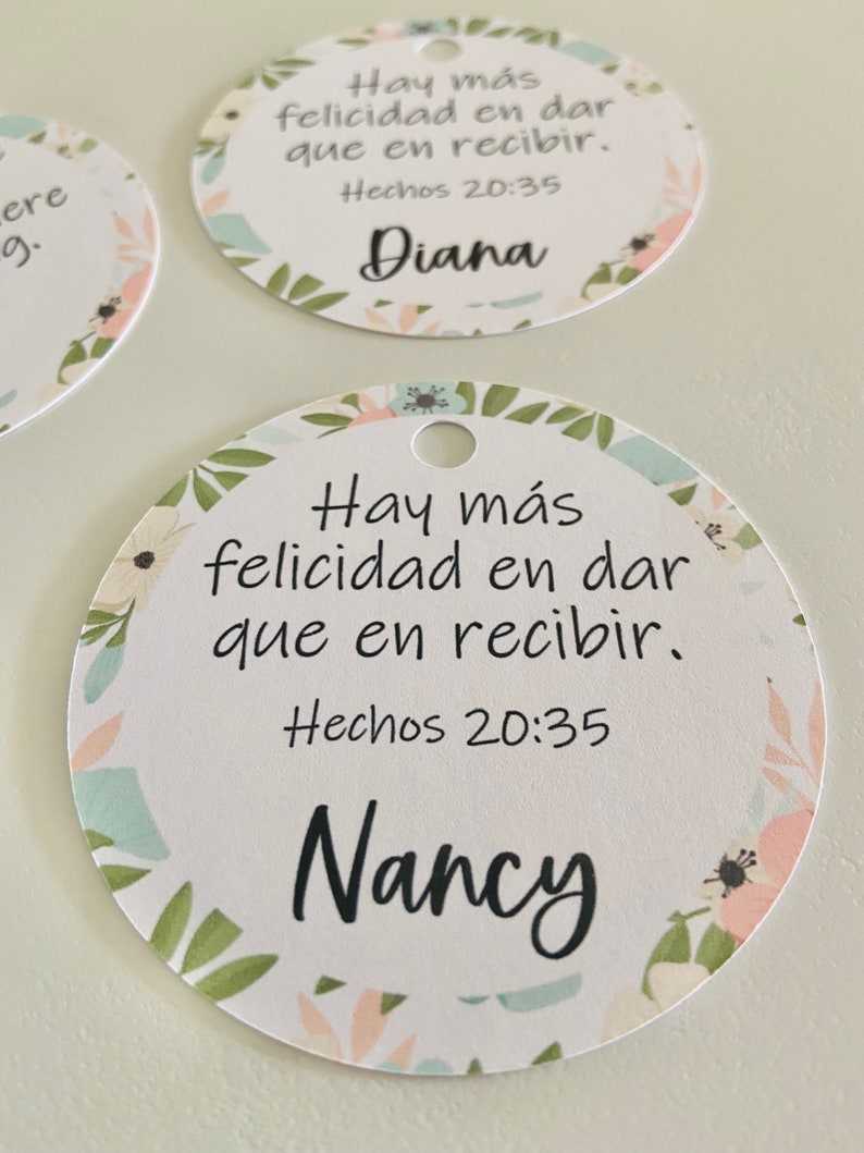 K&ouml;nnte beinhalten: Zwei runde wei&szlig;e Geschenkanh&auml;nger mit Blumenmuster. Die Anh&auml;nger haben oben ein Loch f&uuml;r eine Schnur. Der Text auf den Anh&auml;ngern lautet "Hay m&aacute;s felicidad en dar que en recibir. Hechos 20:35" und die Namen "Diana" und "Nancy".