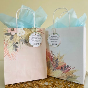 K&ouml;nnte beinhalten: Zwei Geschenkt&uuml;ten mit wei&szlig;en Henkeln und blauem Seidenpapier. Die Taschen haben ein florales Design und ein Zitat &uuml;ber das Gl&uuml;ck des Gebens. Die Taschen sind mit den Namen "Brittney" und "Nancy" beschriftet.