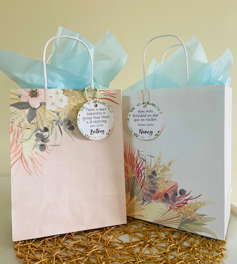 K&ouml;nnte beinhalten: Zwei Geschenkt&uuml;ten mit wei&szlig;en Henkeln und blauem Seidenpapier. Die Taschen haben ein florales Design und ein Zitat &uuml;ber das Gl&uuml;ck des Gebens. Die Taschen sind mit den Namen "Brittney" und "Nancy" beschriftet.