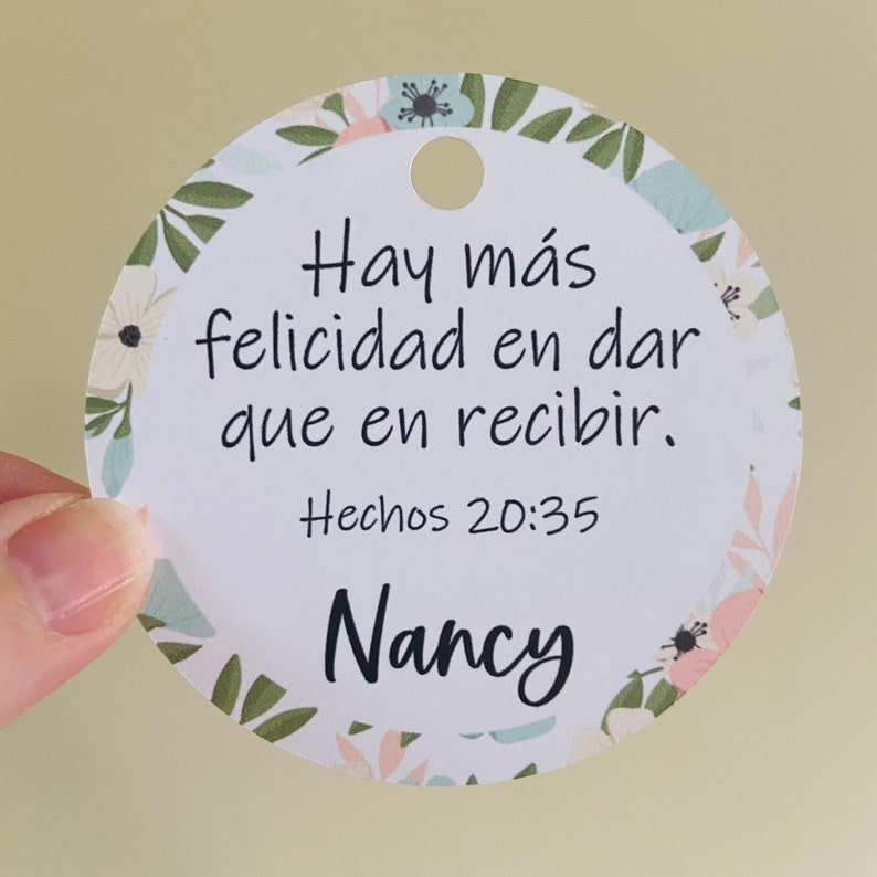 K&ouml;nnte beinhalten: Ein wei&szlig;er runder Anh&auml;nger mit floralem Rand. Der Anh&auml;nger tr&auml;gt den Text "Hay m&aacute;s felicidad en dar que en recibir. Hechos 20:35" und den Namen "Nancy" in Schwarz.