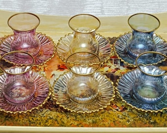 May include: Set of six clear glass teacups with gold rims and matching saucers. The saucers have a scalloped edge and iridescent finish in shades of pink, blue, and gold.