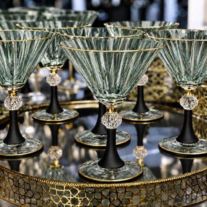 Könnte beinhalten: Ein Set aus Martinigläsern mit salbeigrünen, gerippten Glasschalen und Goldrändern. Jedes Glas hat einen facettierten Kristallkugel-Akzent am Stiel, einen mattschwarzen Sockel und einen goldgeränderten runden Fuß, angeordnet auf einem Goldtablett.