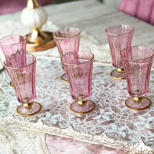 May include: A set of six pink-tinted glass goblets with gold rims and bases. The glasses have a fluted design and are arranged on a decorative tray. The tray has a white floral pattern and a lace edge.