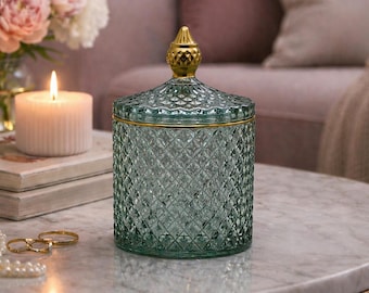 May include: A decorative glass jar with a diamond pattern and a gold-colored lid. The jar is a soft teal color and sits on a marble table. The lid has a gold finial. The jar is a decorative home accent.