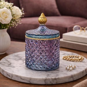 May include: A decorative glass jar with a lid, featuring a diamond pattern. The jar transitions from blue to purple, with gold accents on the lid and rim. It sits on a marble tray, alongside a gold beaded bracelet and a pearl ring.
