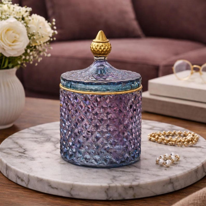 May include: A decorative glass jar with a lid, featuring a diamond pattern. The jar transitions from blue to purple, with gold accents on the lid and rim. It sits on a marble tray, alongside a gold beaded bracelet and a pearl ring.