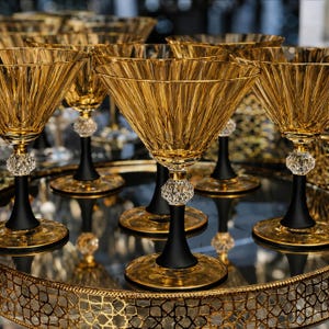 Op de afbeelding: Een collectie amberkleurige glazen coupes met brede, gegroefde kelken. Elk glas heeft een matzwarte steel met een heldere, gefacetteerde kristallen bol direct onder de kelk, rustend op een ronde amberkleurige voet met een goudkleurige rand.