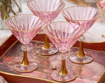 May include: Four pink crystal cocktail glasses with gold rims and stems are arranged on a decorative tray. The glasses have a faceted design and a clear crystal ball detail on the stem. The tray has a floral design.