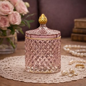 May include: A pink glass jar with a diamond pattern and a gold lid. The jar sits on a white lace doily, with pink roses in the background. A gold ring and pearl earrings are also visible.