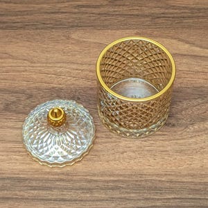 May include: A clear glass jar with a gold rim and diamond pattern, next to its matching lid. The lid has a gold knob. The jar is open, revealing its interior. The items are on a wooden surface.