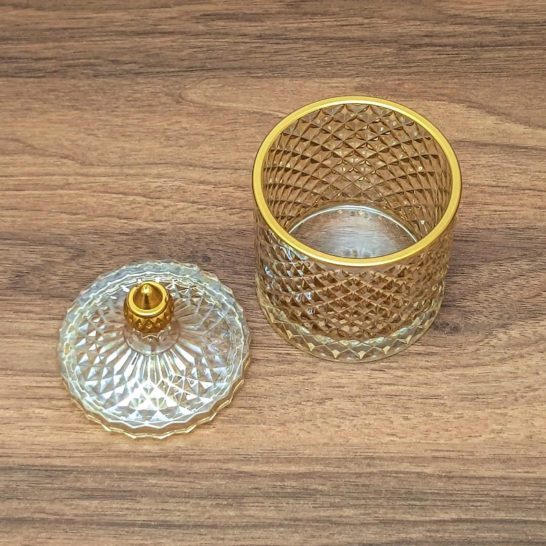 May include: A clear glass jar with a gold rim and diamond pattern, next to its matching lid. The lid has a gold knob. The jar is open, revealing its interior. The items are on a wooden surface.