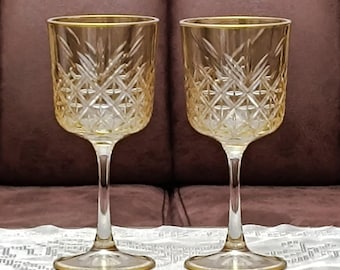 Könnte beinhalten: Zwei bernsteinfarbene Weingläser mit einem Diamantschliffmuster. Die Gläser haben einen goldenen Rand und einen langen, schlanken Stiel. Die Gläser stehen vor einem braunen Hintergrund.