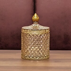 May include: A decorative jar with a diamond pattern and gold-coloured lid and trim. The jar is made of clear glass and sits on a wooden surface. The lid has a small, gold-coloured knob.
