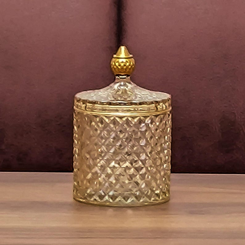 May include: A decorative jar with a diamond pattern and gold-coloured lid and trim. The jar is made of clear glass and sits on a wooden surface. The lid has a small, gold-coloured knob.