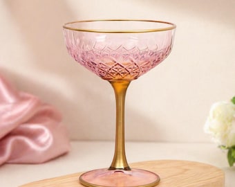 May include: A pink champagne coupe with a gold rim and stem. The glass has a diamond pattern on the bowl and a circular base. The coupe is set on a wooden board, with a pink cloth in the background.