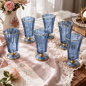 Könnte beinhalten: Ein Set aus sechs blauen Glasbechern mit vertikal gerippten Texturen, goldfarbenen Rändern und goldfarbenen runden Sockeln. Jeder Becher verfügt über einen schlanken Stiel, der die geriffelte Schale mit dem Sockel verbindet.