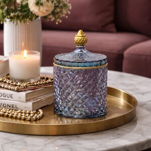 May include: A decorative glass jar with a lid, featuring a diamond-cut pattern in shades of blue and purple. The jar has a gold rim and a gold-colored finial on the lid. It sits on a gold tray, alongside a lit candle and a stack of books.