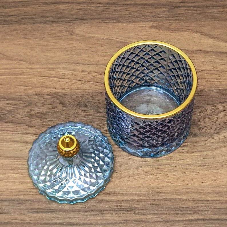 May include: A cylindrical glass jar with a blue diamond-cut pattern and a gold-rimmed opening. The matching circular lid features a matching diamond-cut texture and a small, textured gold-colored knob handle at the center.
