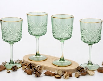 May include: Four green wine glasses with gold rims and diamond-patterned detailing. The glasses sit on a wooden oval tray, surrounded by decorative dried potpourri. The stemware is ideal for serving wine or other beverages.