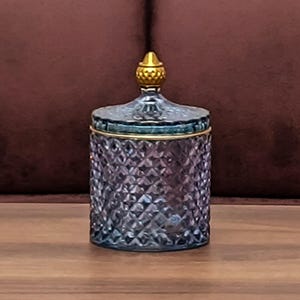 May include: A cylindrical glass jar with a purple-tinted, diamond-cut textured surface. The jar features a matching lid topped with a small, gold-colored, textured finial and a thin gold-colored rim around the top edge of the container.