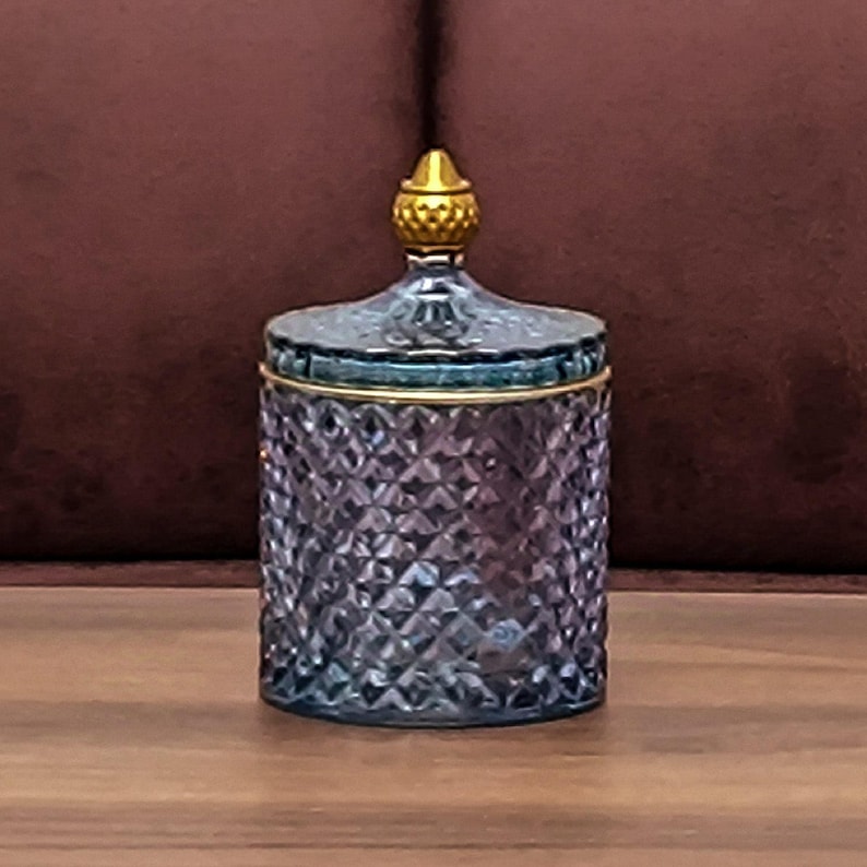 May include: A cylindrical glass jar with a purple-tinted, diamond-cut textured surface. The jar features a matching lid topped with a small, gold-colored, textured finial and a thin gold-colored rim around the top edge of the container.