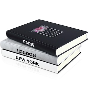Könnte beinhalten: Drei Hardcover-Bücher, die übereinander gestapelt sind. Das oberste Buch ist schwarz mit einem Blumendesign und dem Text "France" auf dem Cover. Das zweite Buch ist weiß mit schwarzem Text, der "Paris" sagt. Das unterste Buch ist weiß mit schwarzem Text, der "London" und "New York" sagt.