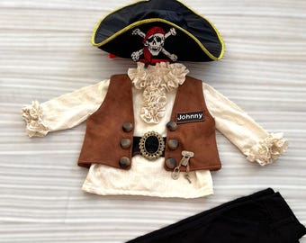May include: A pirate costume set featuring a black hat with a skull and crossbones, a brown vest with large buttons, a white shirt with ruffled accents, black pants, and a belt. The name "Johnny" is on the vest.