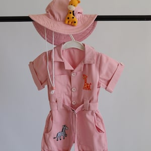 May include: A pink baby jumpsuit with short sleeves and a belt. The jumpsuit has two embroidered giraffes and a zebra. The jumpsuit is paired with a pink sun hat with an embroidered giraffe.