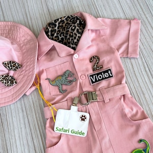 May include: Pink safari jumpsuit with a dinosaur patch, a leopard print scarf, a hat, and a name tag that says "Safari Guide". The jumpsuit has a belt and a patch that says "Violet 2".