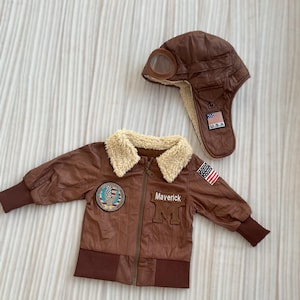 May include: A brown bomber jacket and aviator hat set. The jacket has a faux fur collar, the word "Maverick" and the letter "M" on the front, and a U.S.A. flag patch. The hat has goggles and a U.S.A. flag patch.
