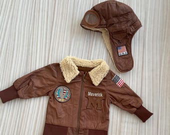 Könnte beinhalten: Braune Bomberjacke und Fliegermützen-Set. Die Jacke hat einen Kunstfellkragen, das Wort "Maverick" und den Buchstaben "M" auf der Vorderseite sowie einen U.S.A.-Flaggenaufnäher. Die Mütze hat eine Brille und einen U.S.A.-Flaggenaufnäher.