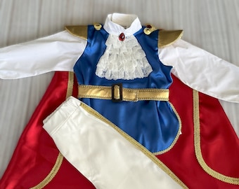 May include: A red cape with gold trim, a blue waistcoat with gold trim and a white shirt, and white trousers. The waistcoat has a white lace collar and a gold belt.