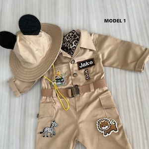 Puede incluir: Un mono de safari de color beige con una cebra en blanco y negro, un león marrón y un parche de un jeep verde. El mono tiene un cinturón marrón con una cuerda amarilla atada. El atuendo también incluye un sombrero de color beige con orejas de Mickey Mouse negras y una bufanda de leopardo marrón y negro.