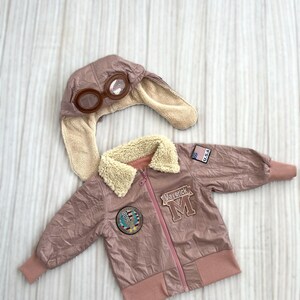 May include: A dusty rose-colored aviator jacket and matching hat with goggles. The jacket has a cream-colored faux fur collar, a zipper closure, and patches that read "Maverick" and "U.S.A.". The hat has ear flaps and clear goggles.