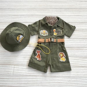 May include: A green safari-themed outfit for a child, featuring a hat, a short-sleeved jumpsuit with pockets, a belt, and a yellow lanyard. The jumpsuit has patches with animal illustrations and the text "Safari" and "Sevyyn".